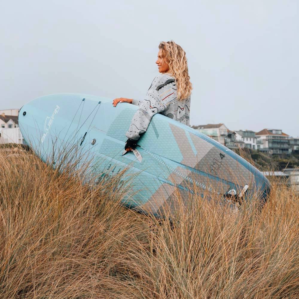 Woman with curly hair holding a light blue paddleboard in tall grass near the beach.