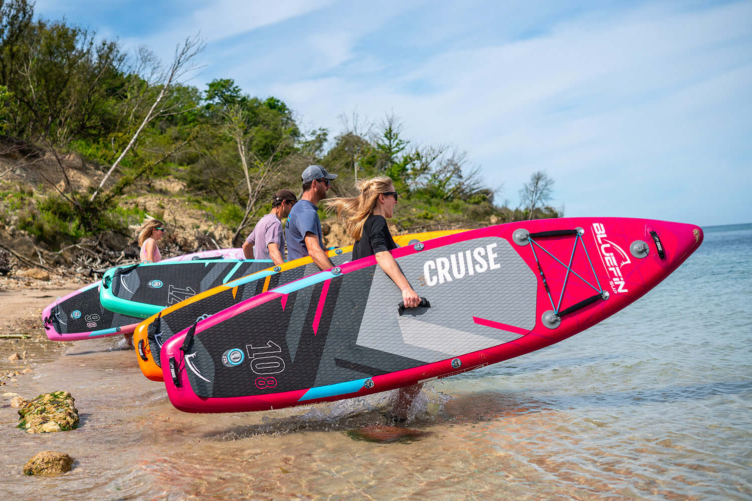 Three people carrying colorful inflatable paddleboards along a beach with water and trees in the background.