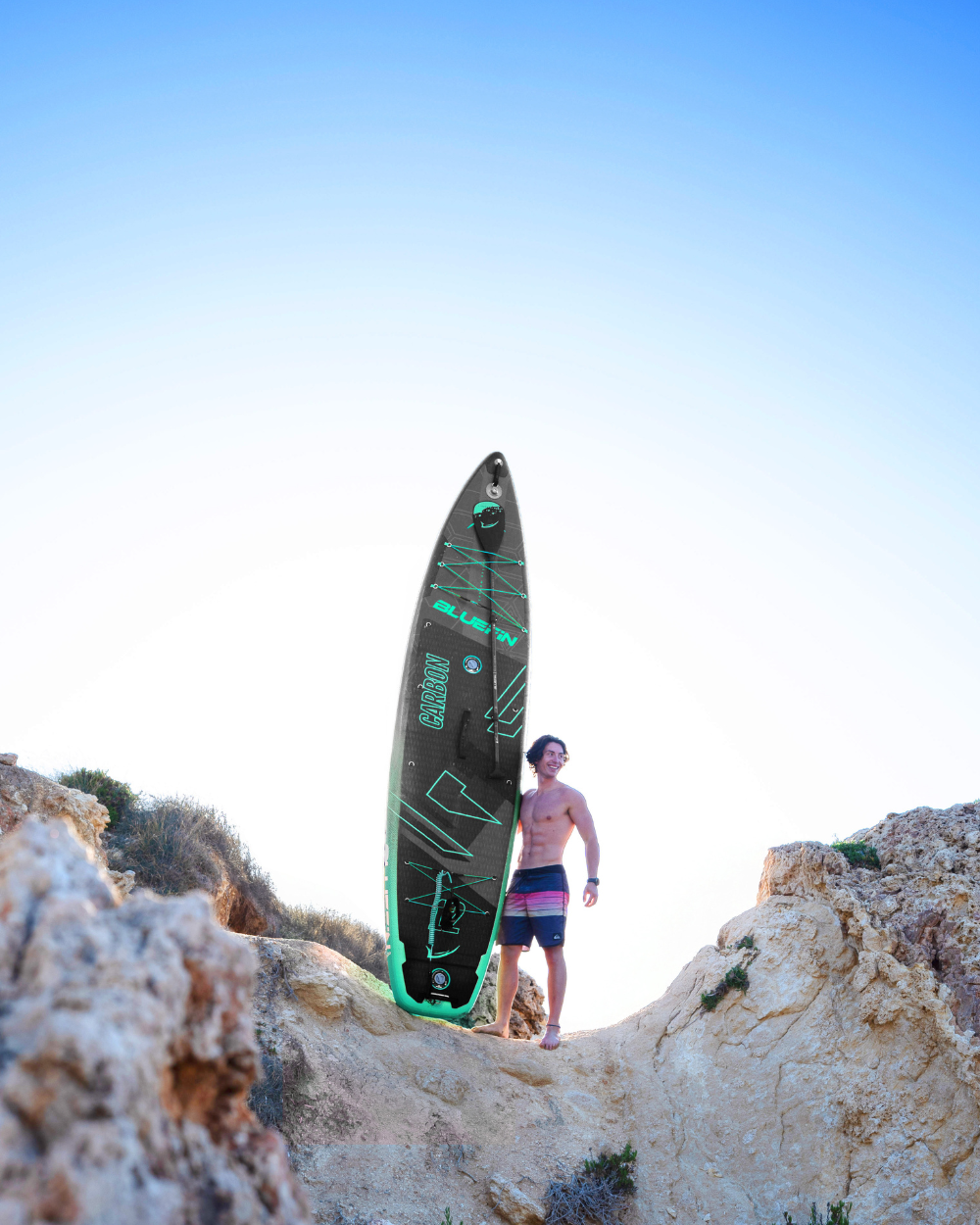 Man standing on rocks holding a black inflatable paddleboard with green accents and textured deck pad