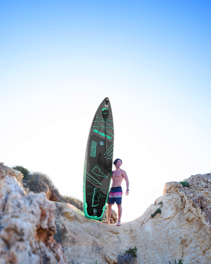 Man standing on rocks holding a black inflatable paddleboard with green accents and textured deck pad