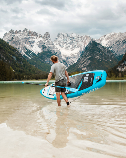 Person walking in shallow water carrying a blue inflatable paddleboard with mountains in the background