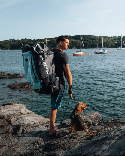 Man with a black and teal backpack standing on rocks by the water with a dog on a leash.