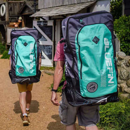 Two people walking with large Bluefin Carbon SUP backpacks on a gravel path near buildings.