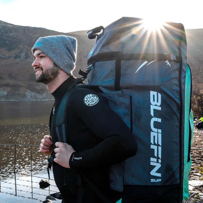 Man in wetsuit carrying a large Bluefin SUP backpack by a lake with mountains in the background.