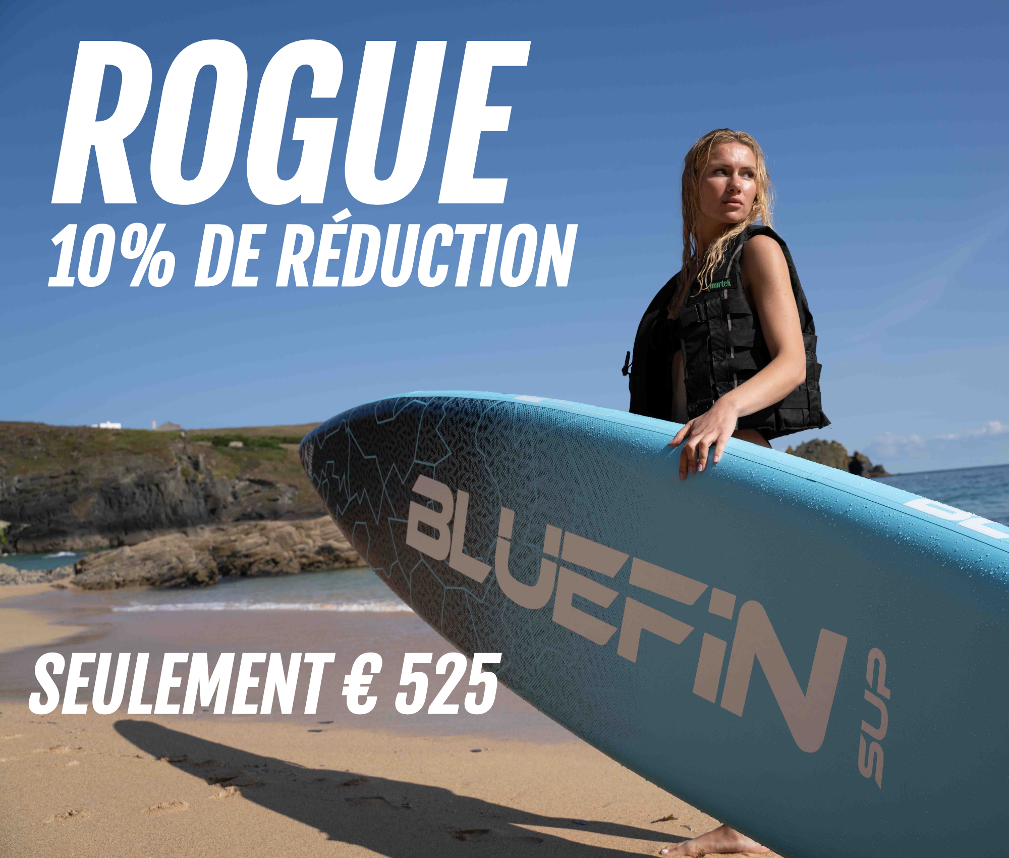 Woman holding a blue Bluefin SUP paddleboard on a beach with rocky background and clear sky