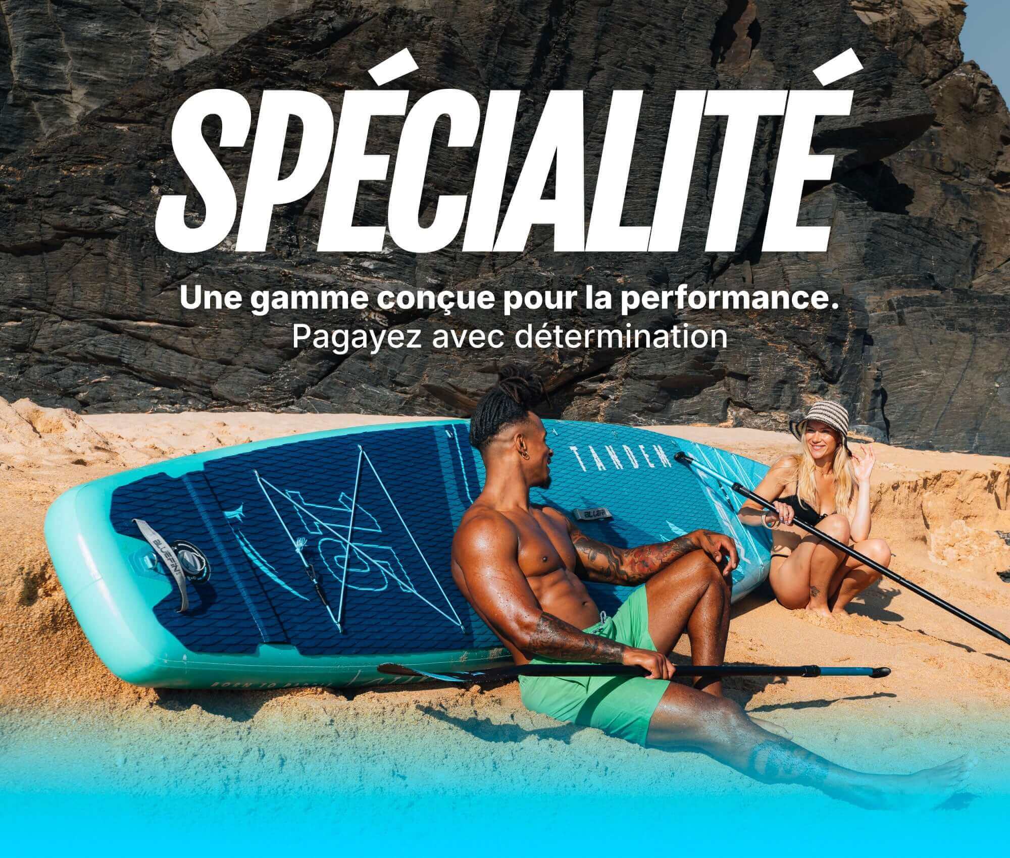 Two people on a beach with an inflatable paddleboard, one sitting and one waving, both in swimwear.