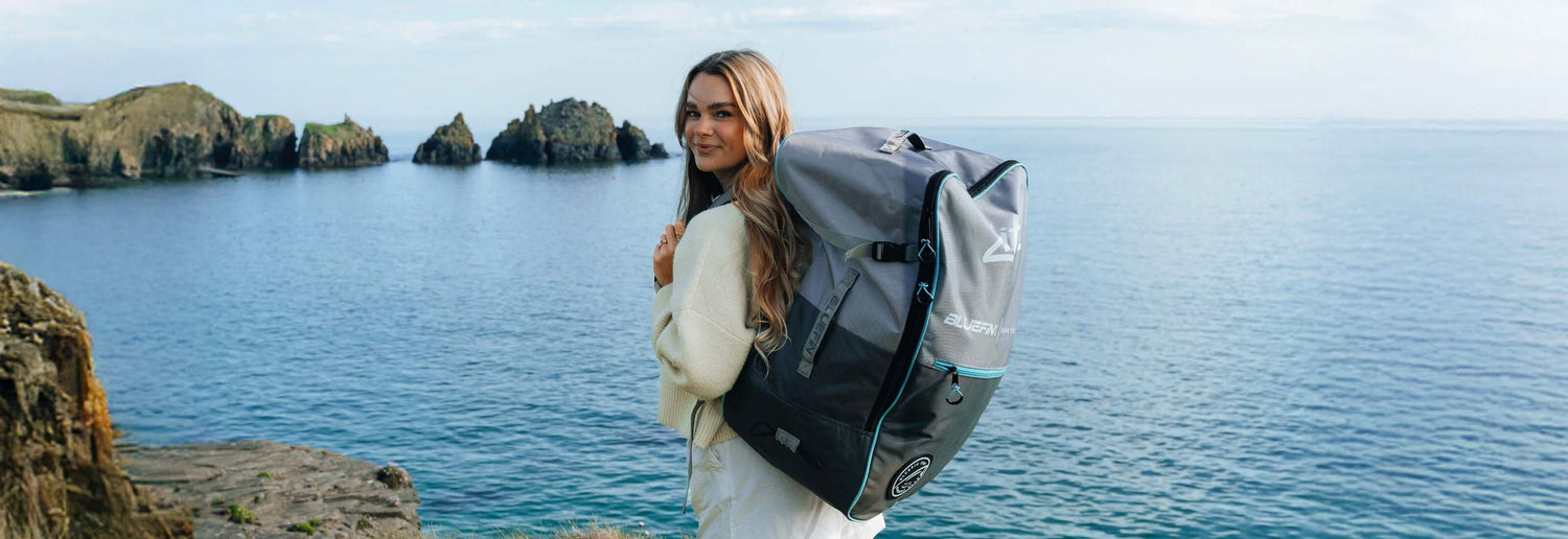 Woman wearing a white sweater, carrying a large gray Bluefin SUP backpack by the water.