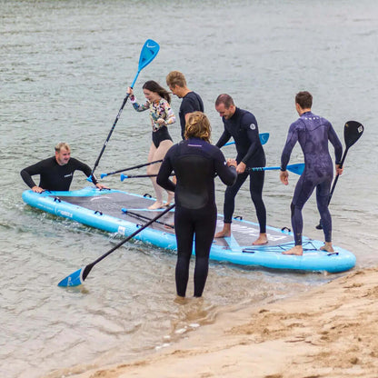 Groupe de six personnes sur un grand paddleboard gonflable bleu dans une eau peu profonde, certains pagayant, d'autres en équilibre.
