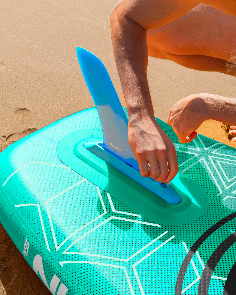 Person attaching a blue fin to a green inflatable paddleboard on sandy beach