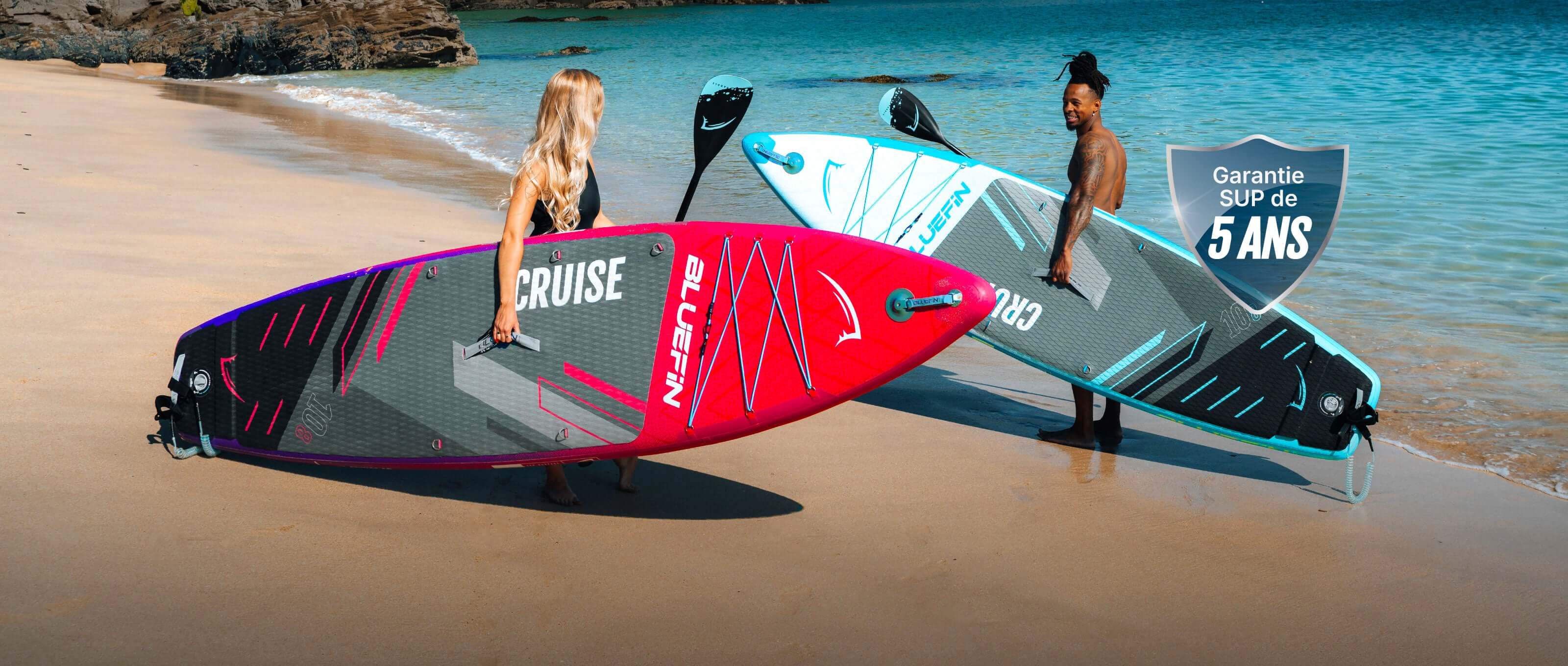 Two inflatable paddleboards on a beach, one pink and one blue, with two people holding them.