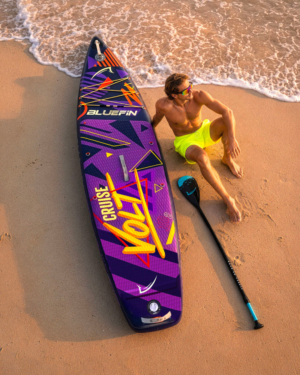 Inflatable paddleboard with colorful design, man in yellow shorts sitting on sand, paddle nearby