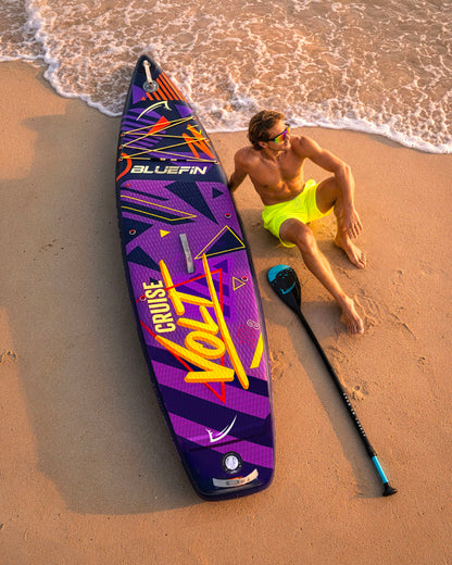Inflatable paddleboard with colorful design, man in yellow shorts sitting on sand, paddle nearby