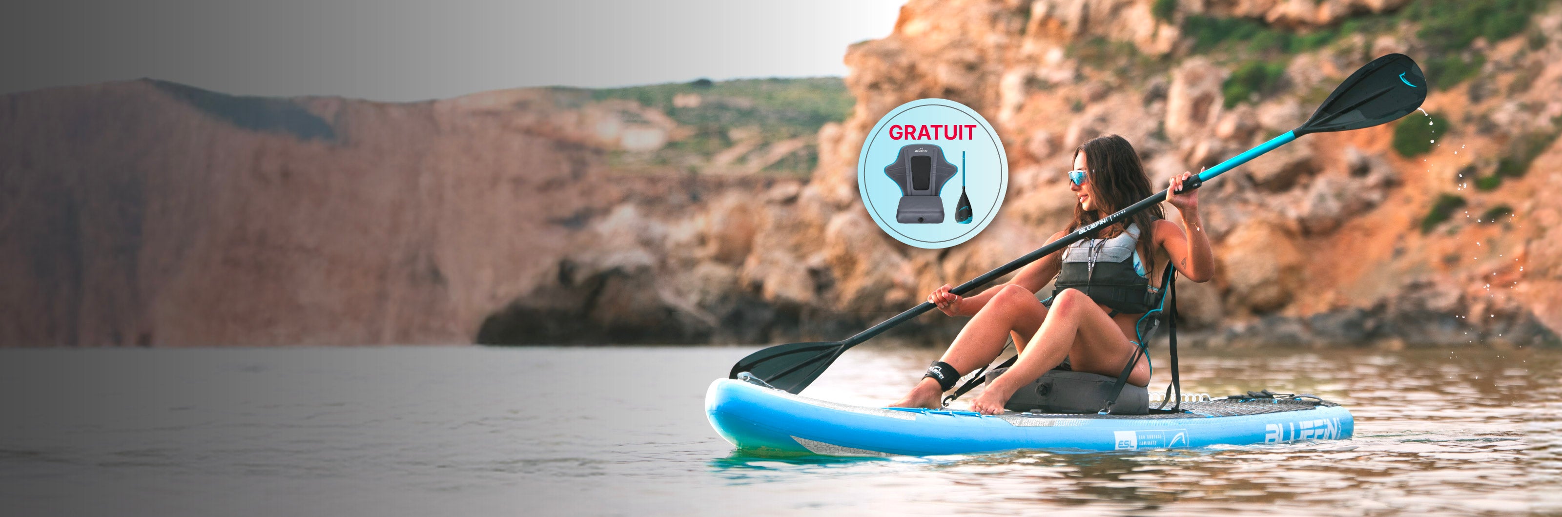 Woman paddling on a blue inflatable paddleboard in calm water, rocky cliffs in background